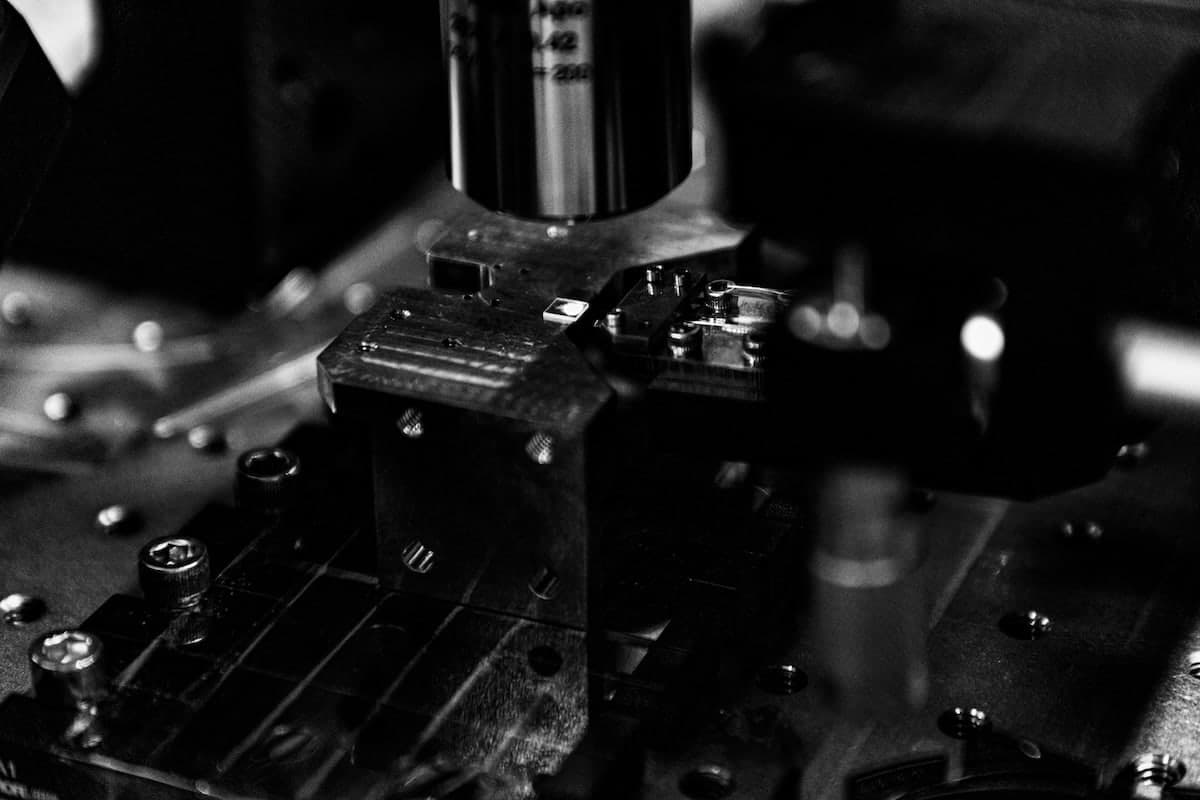 A close-up, black-and-white photograph of a diamond photonic integrated circuit (PIC) under a microscope in the Lightsynq lab. The circuit is mounted on a precision stage with intricate components and structures visible.
