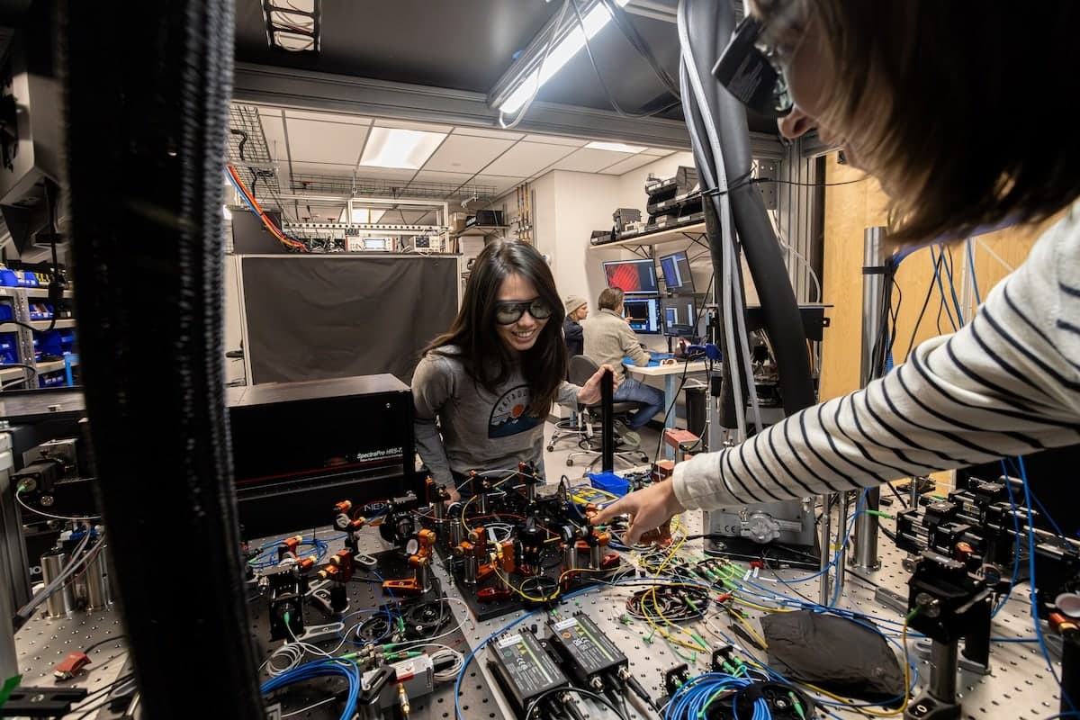 A research team working together in Lightsynq's quantum interconnect lab at their Boston, MA headquarters. The lab features complex optical and cryogenic test equipment on a table, with multiple cables and laser components.